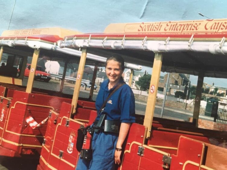 Image shows: Hilary Roberts, posing in front of the Abroath tourist land train, where she worked as a conductor in 1994 and 1995.Hilary is dressed in navy trousers and t-shirt and is wearing her conductor's belt and ticket machine.