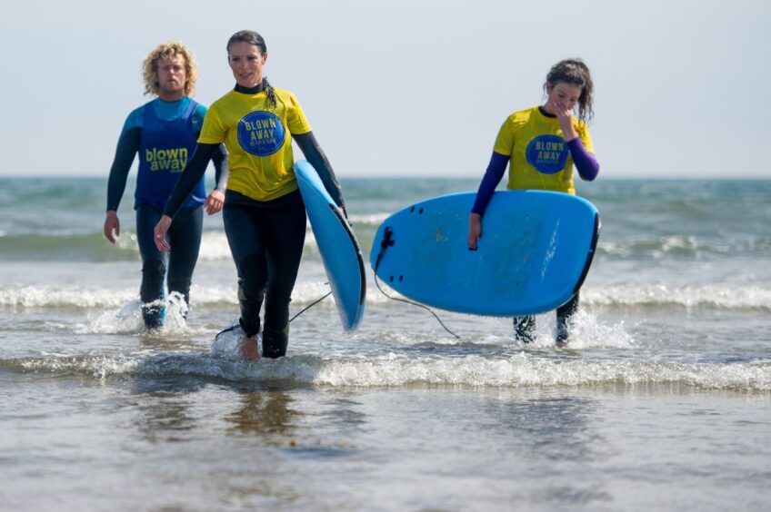 Image shows: A surf lesson with Blown Away at West Sands beach in St Andrews. Two surfers wearing wetsuits and yellow t-shirts are in the foreground carrying blue surf boards. The is a watersports instructor wearing a blue t-shirt and wetsuit.