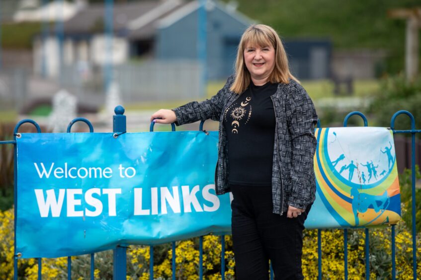 Image shows: Hilary (she/her) Roberts who had a summer job on the Abroath Tourist Train. Hilary is pictured standing beside a sign that says Welcome to West Links in Arbroath. She is wearing black trousers and t-shirt with a grey cardigan and had shoulder length brown hair with a fringe.