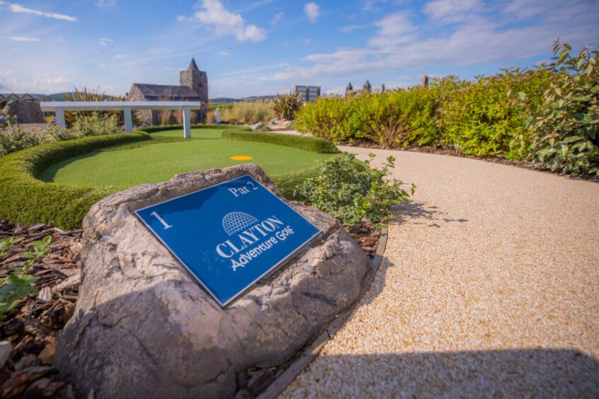 Image shows: the mini golf course at Clayton Caravan Park near St Andrews. There is a sign for the first hole on the course and some of the St Andrews themed landmarks in the background, the photo is taken on a bright, sunny day.