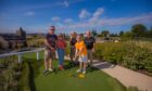 Image shows: A happy family getting ready to play mini golf at Clayton Adventure Golf. There is a family on holiday standing around one of the tees at the mini golf course. A boy holding a golf club at the front of the picture is wearing a yellow t-shirt and white shorts is smiling broadly at the camera.