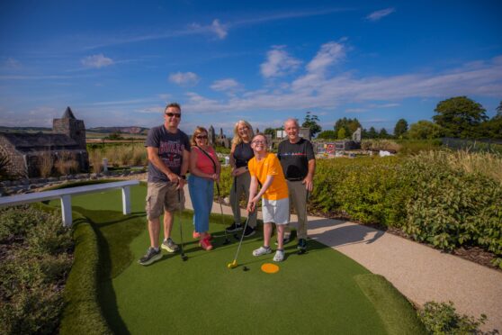 Image shows: A happy family getting ready to play mini golf at Clayton Adventure Golf. There is a family on holiday standing around one of the tees at the mini golf course. A boy holding a golf club at the front of the picture is wearing a yellow t-shirt and white shorts is smiling broadly at the camera.