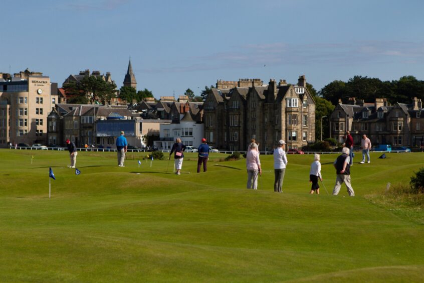 Image shows: A wide view of the Himalayas putting green in St Andrews. The course is busy with people practising their putting skills and the town of St Andrews is in the background.