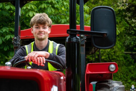 Image shows: Elliot Comerford who has a summer job at Craigtoun Park in St Andrews. Elliot is sitting in the driver's seat of the red tractor called Puffing Billy. He is wearing a high vis vest and black jumper.