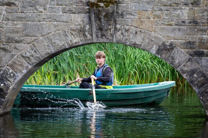 Image shows: Elliot Comerford who has a summer job at Craigtoun Country Park in Fife. Elliot is in a green rowing boat, which is beside an arched bridge.