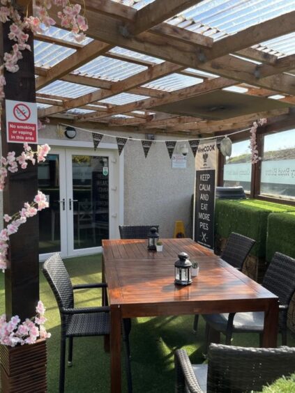 An Angus al fresco dining spot, The WeeCOOK Kitchen, as shown, with a wooden table, black chairs and a canopy overhead with floral decoration. 