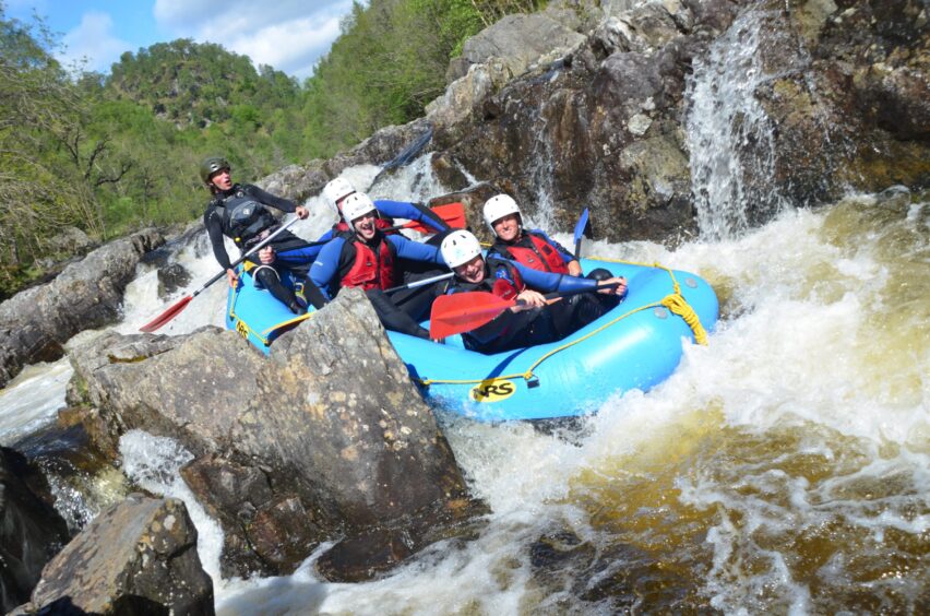 Image shows: an inflatable raft on the River Tummel. The raft is full of people negotiating the white water of the river, with an instructor keeping everyone safe at the back of the raft. Family watersports fun.