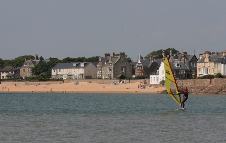 Image shows: A windsurfer in action at the beach in Elie. The colourful yellow sail of the windsurf board cuts across the calm water with the village of Elie and its sandy beach in the background.