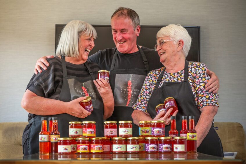 Allan stands with his wife, Joan, and her mother, Betty, smiling and laughing. On the table in front of them are chilli sauces and chilli jellies. 