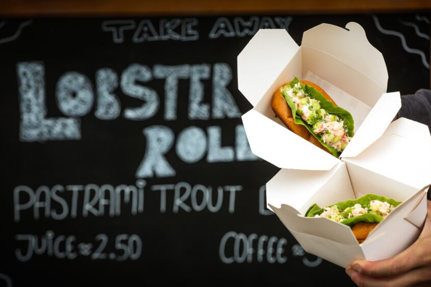 Two lobster rolls (a bun with lettuce and a creamy lobster filling) held out in front of a blackboard sign. 