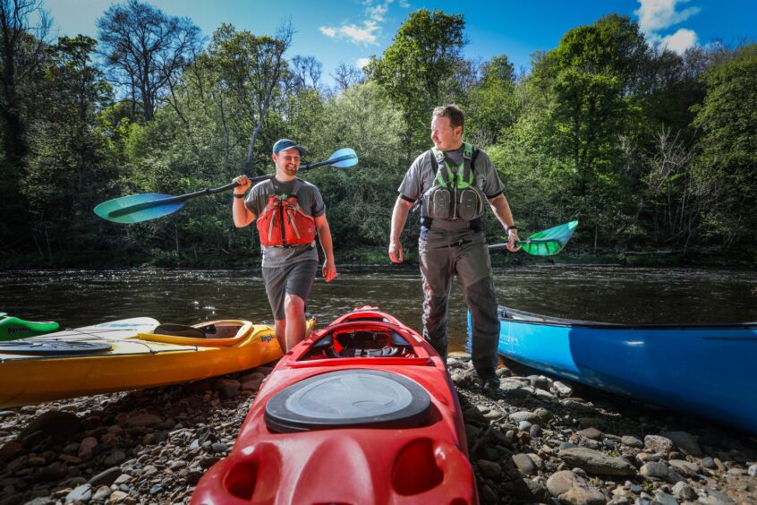 Image shows: James Barrett and Danny Boyd of Outdoor Explore. The two men are holding kayak paddles and are standing on the riverbank among brightly coloured kayaks. Family watersports feature.