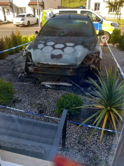 Charred car in driveway with police van behind