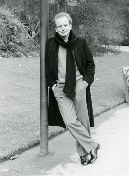 McDiarmid, in long coat and scarf, looks at the camera while leaning against a lamppost