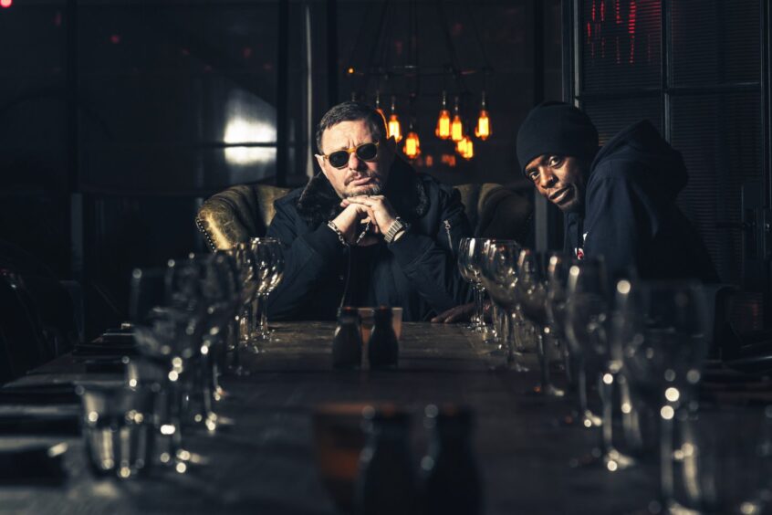 Shaun Ryder and Kermit of Black Grape at the end of a long banqueting table set with glasses and plates