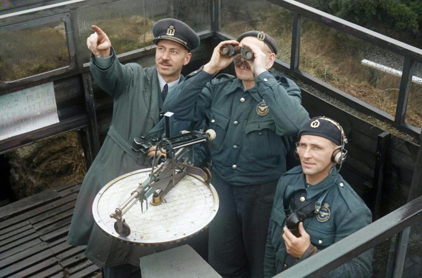 Three men in uniform in an observation tower, with one using binoculars and one wearing a headset, while the third points to the sky