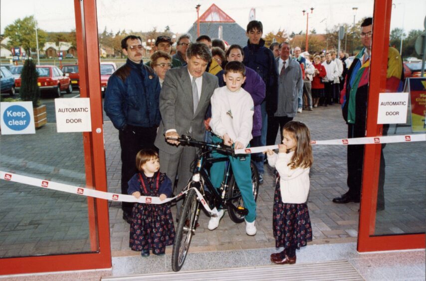 Kevin Loach won a mountain bike when the new store opened in 1993. He sits on it to cut the ribbon as shoppers queue behind him