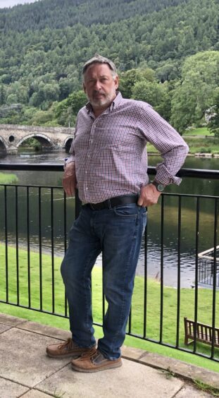 Colin Morton in jeans and checked shirt leaning against railings beside River Tay at Kenmore