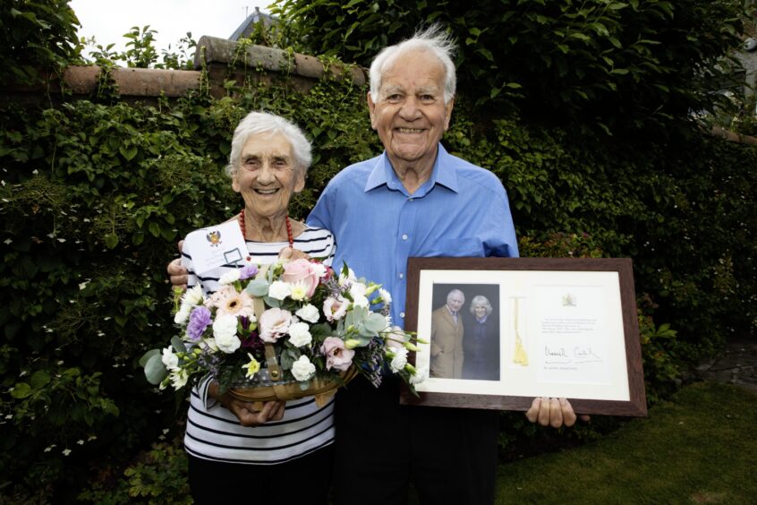 Mabel and Eddie Hartley in garden holding basket of flowers and framed card from King Charles and Queen Camilla.