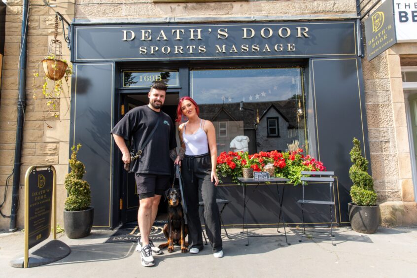 James and Lauren standing outside black painted Death's Door Sports Massage shopfront with five month-old Doberman puppy