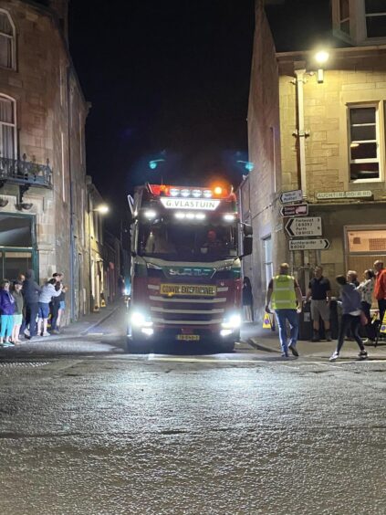 Lorry reversing up narrow street in centre of Dunning watched by large late night crowd