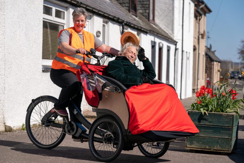 Smiling Rowen Ross piloting a trishaw with her father seated in front and lifting his hat for the camera