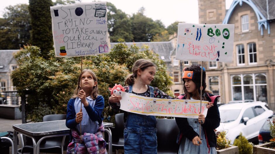 Three little girls holding handmade 'Save Our Library' banners outside Birnam Library.