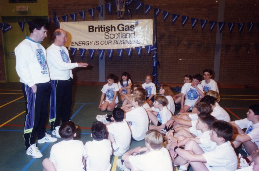 Sir Bobby and a member of his coaching team speak to a group of soccer school attendees in a gym hall