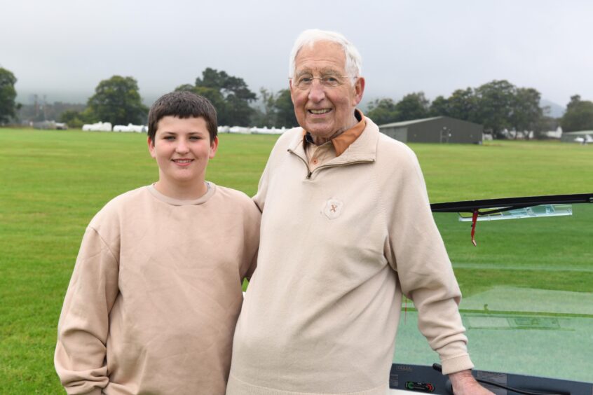 Image shows: Tom Docherty, 90, gliding enthusiast and his grandson Thomas Docherty, 13. Both are wearing beige coloured jumpers and are smiling at the camera. They are at Portmoak Airfield with green grass and trees in the background.