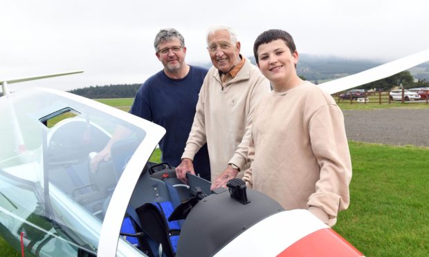 Image shows: Three men standing by the cockpit of a glider. The glass roof of the glider is open and the runway and misty hills are visible in the backgroups. Pictured are: From left to right, Adrian Docherty, Tom Docherty and Charlie Docherty.