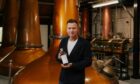 Ewan McGregor holding bottle of whisky in front of large copper still in distillery