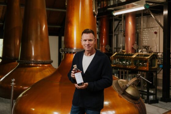 Ewan McGregor holding bottle of whisky in front of large copper still in distillery