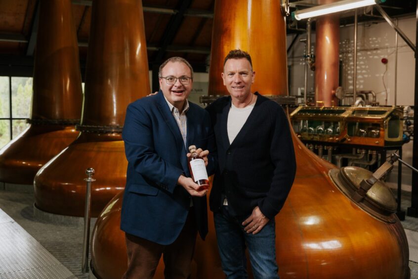 Ewan McGregor and CHAS CEO Rami Okasha holding bottle of whisky in front of copper still at Lochranza distillery