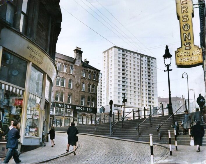 a colourised photo showing people walking on the Wellgate steps in Dundee with shops nearby and a high-rise building in the distance