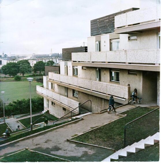 children play on bikes on path beside Dundee's Whitfield blocks, with a view of a field, some trees and buildings in the distance