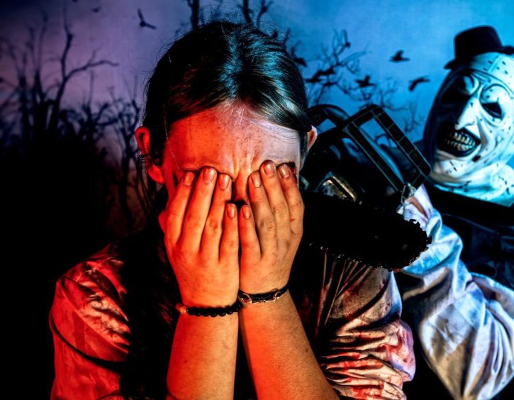 Image shows: a woman with her hands covering her face being menaced by a terrifying Halloween figure wielding a chainsaw.