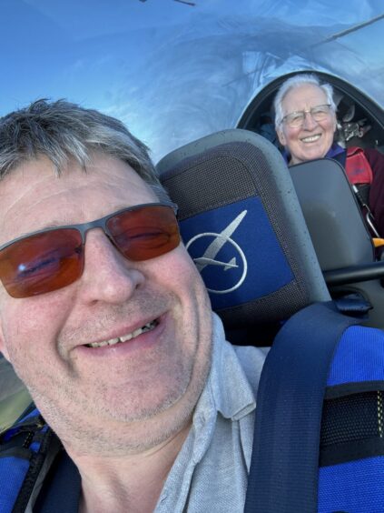 Image shows a close up shot of two men in the cockpit of a glider. Adrian Docherty is in the front seat, he has grey hair and is wearing sunglasses and a wide smile. Tom Docherty is in the back seat also grinning widely.