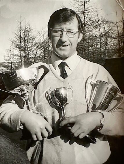 Image shows: A black and white photograph of Tom Docherty showing off some of his gliding trophies. Tom has dark hair and is wearing glasses, a shirt and tie and v-neck jumper.