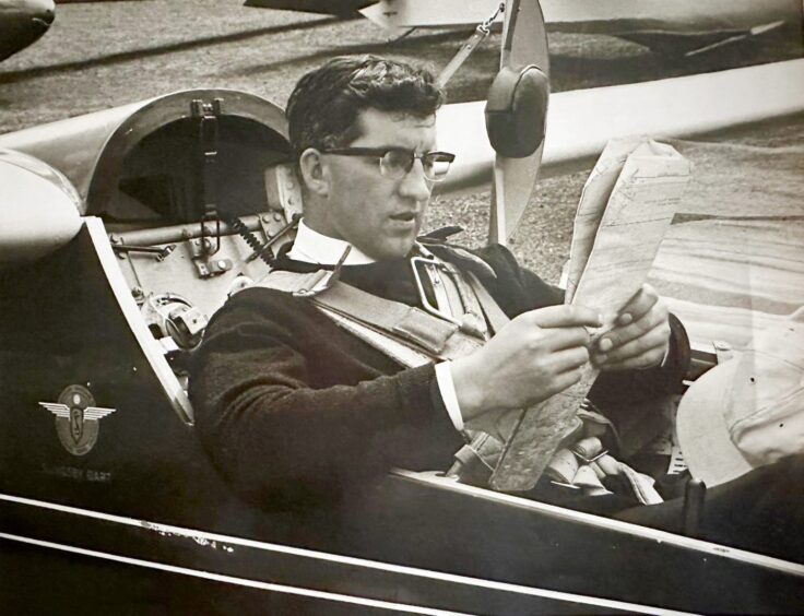 Image shows: A back and white image of a young Tom Docherty. Tom is wearing glasses and is sitting in the cockpit of a glider with a map in his hands.