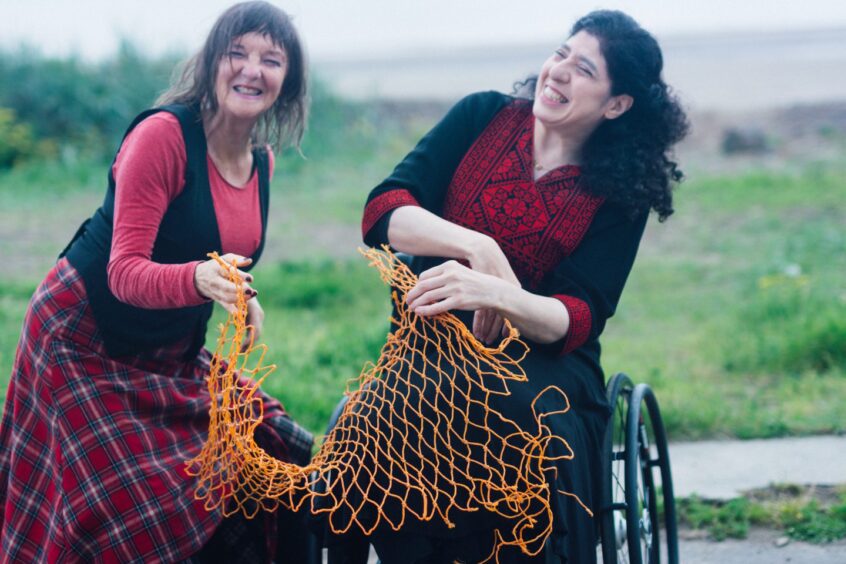 Janis and Nada of Mending Nets. In a field they are laughing with each other and holding an orange net. 