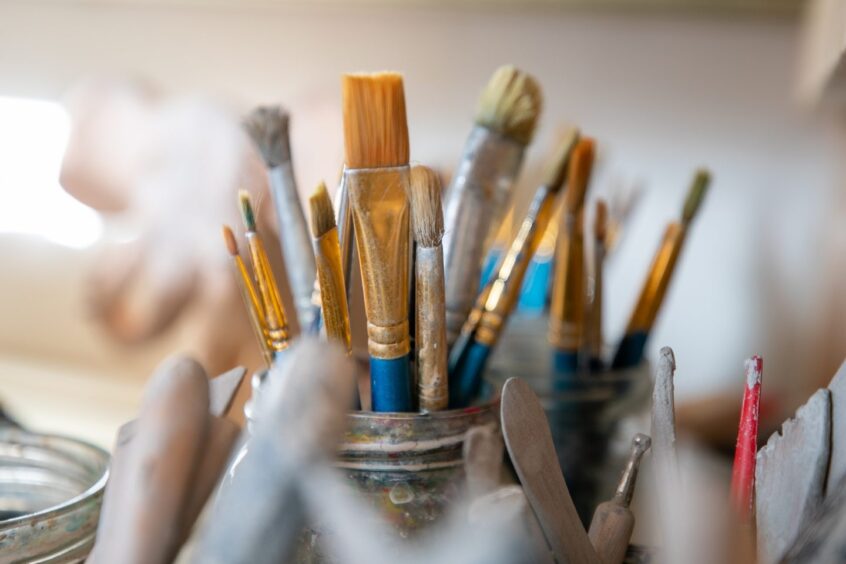 Image shows: A close up shot of a pot with paintbrushes in a pottery workshop. In the foreground, there are some of the tools used to shape and mould clay. Taken at The Workshop in Perth to illustrate a kids party feature.