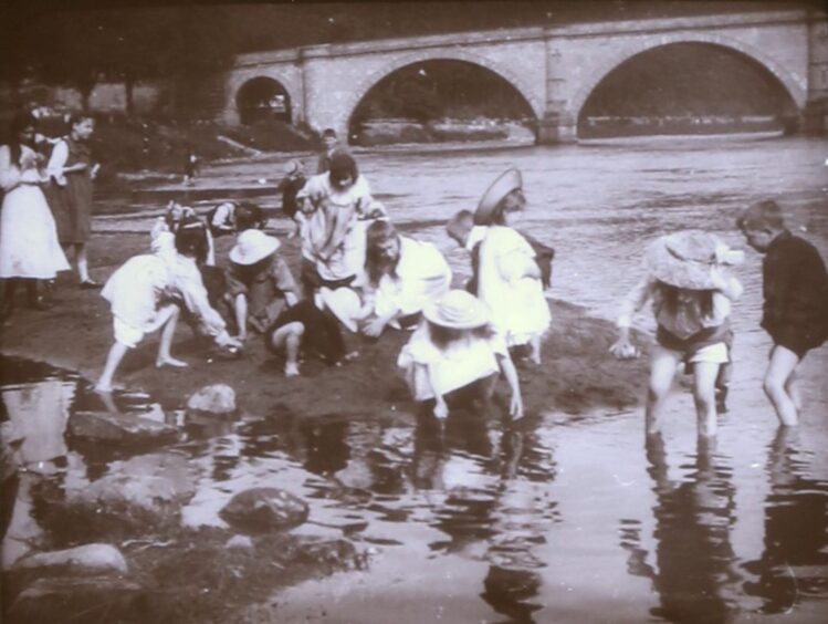 Children in Victorian clothes playing in river in front of Dunkeld bridge.