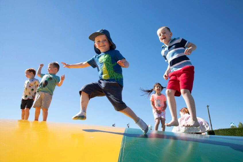 Image shows: a group of happy children jumping on a colourful outdoor inflatable pillow. There is a blue sky in the background and the children are wearing brightly coloured clothes and smiling. The jumping pillow is at Active Kids in Stanley which offers kids birthday parties.