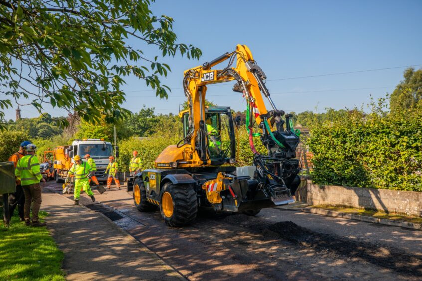 Pothole fixing machine going along road, with workers around it