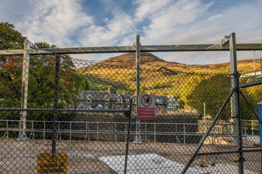 View looking through metal fence beyond structure to St Fillans residents homes and hill behind.