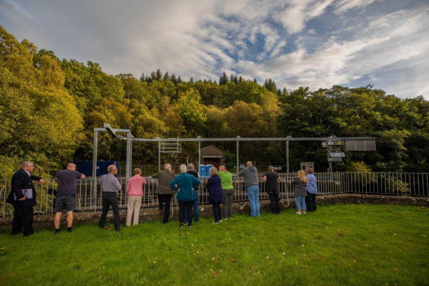 People leaning over fence looking at tall metal structure with trees behind