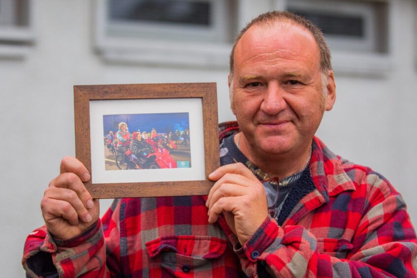 Ian Ross holding framed photo of Rowen Ross piloting a trishaw with two older people seated in front at an evening event