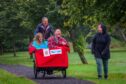 Two people laughing seated in trishaw, as man pilots it through Auchterarder park and young woman walks alongside laughing