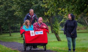 Two people laughing seated in trishaw, as man pilots it through Auchterarder park and young woman walks alongside laughing