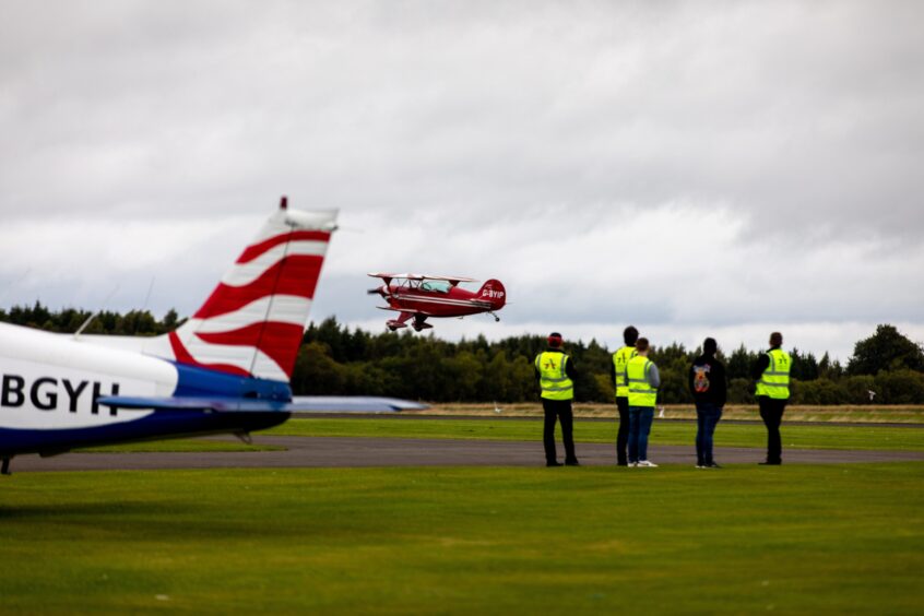 Small red stunt plane coming in to land at Perth airport runway watched by small group of people