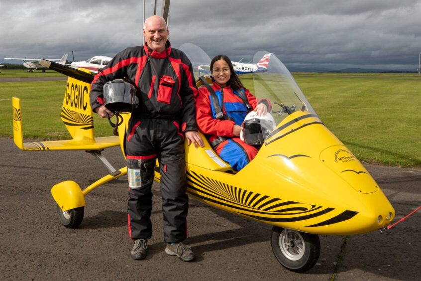 Man standing next to yellow gyrocopter on runway with woman in seat